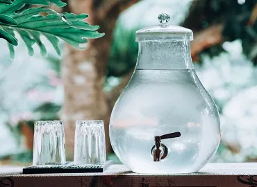 A glass water jug with spigot in a hotel lobby with glasses next to it for hotel guests to enjoy.